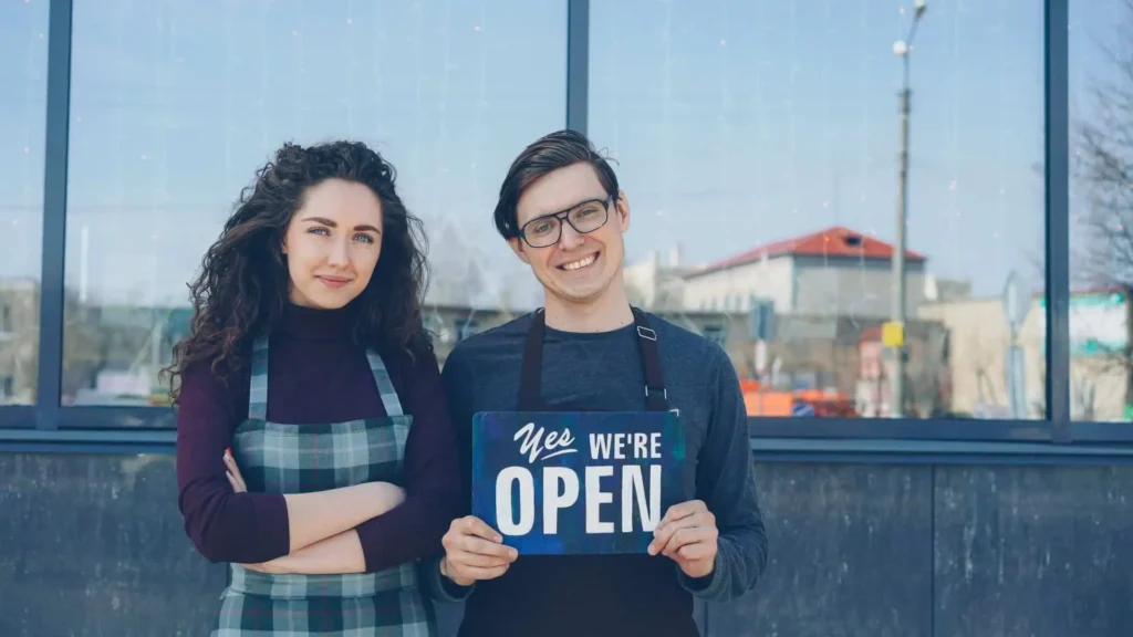 Two small business owners standing outside their shop with an open sign.