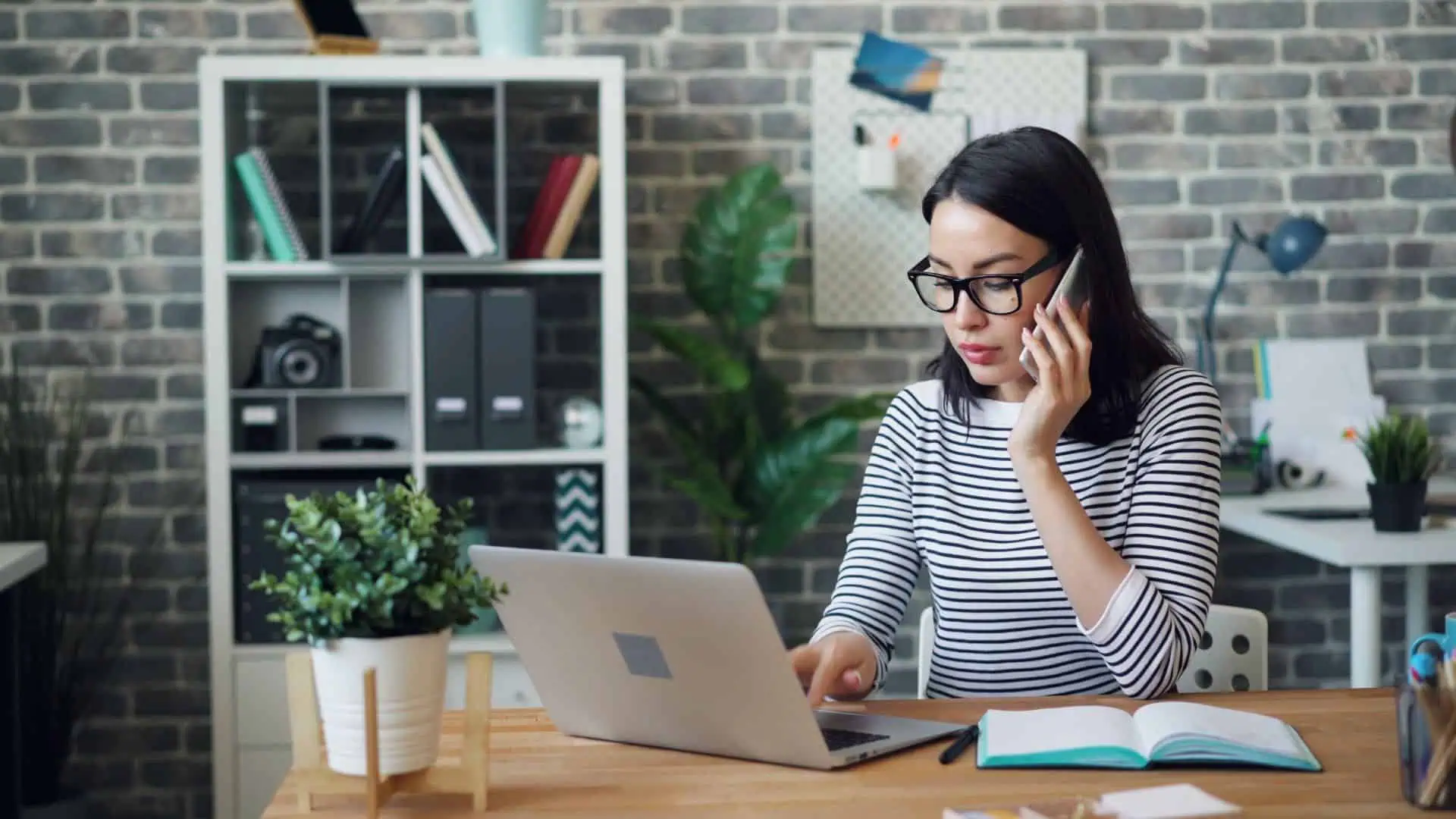 Woman talking on the phone while working on a laptop at an office desk.
